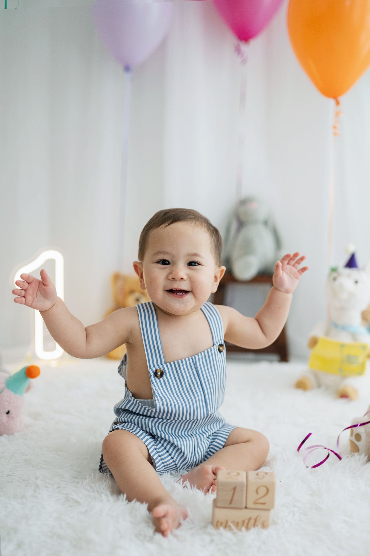 Smiling baby in striped romper sits on white rug surrounded by colorful balloons and toys at a first birthday party.