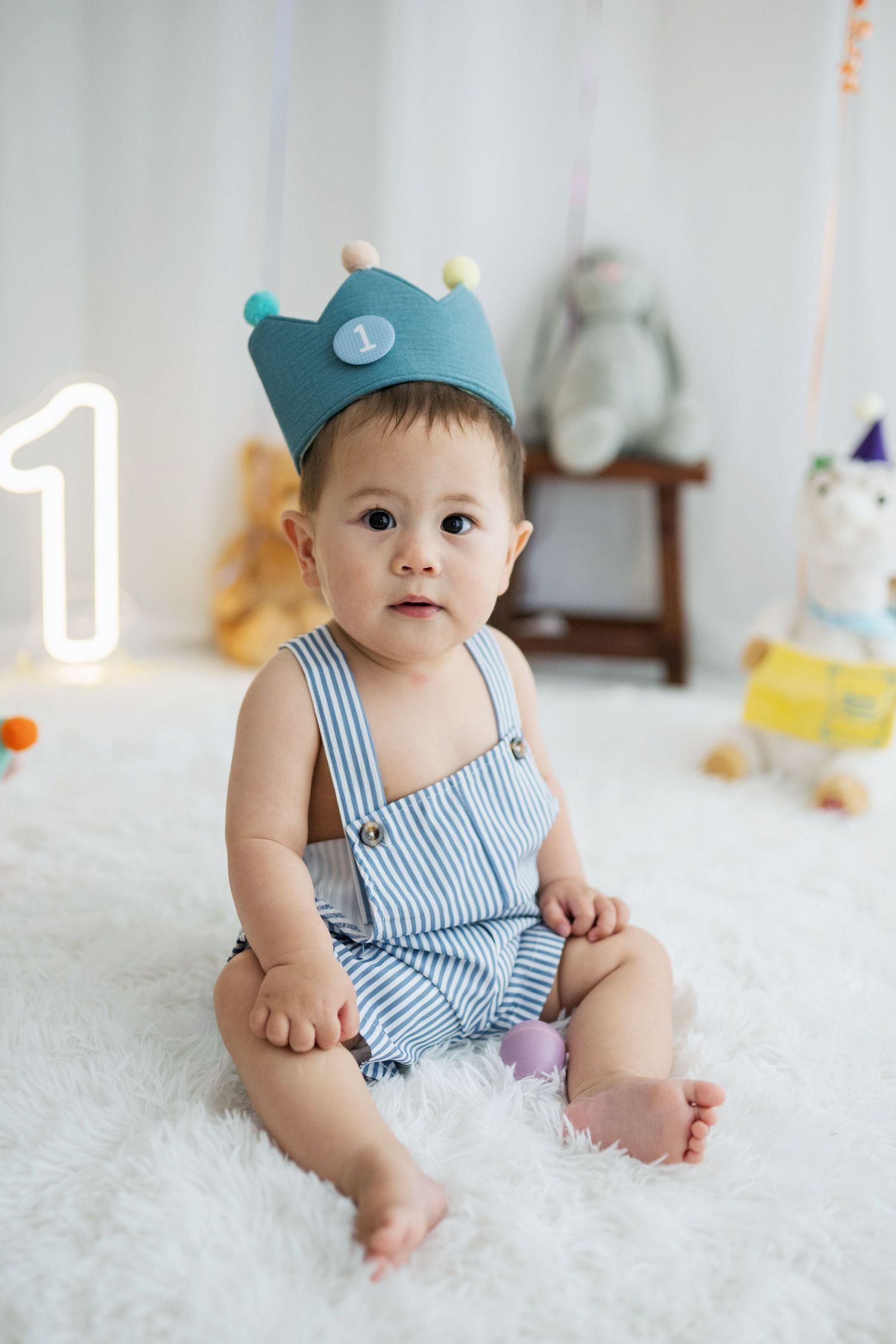 Adorable baby in striped overalls and blue crown hat sits on white fluffy rug celebrating first birthday.