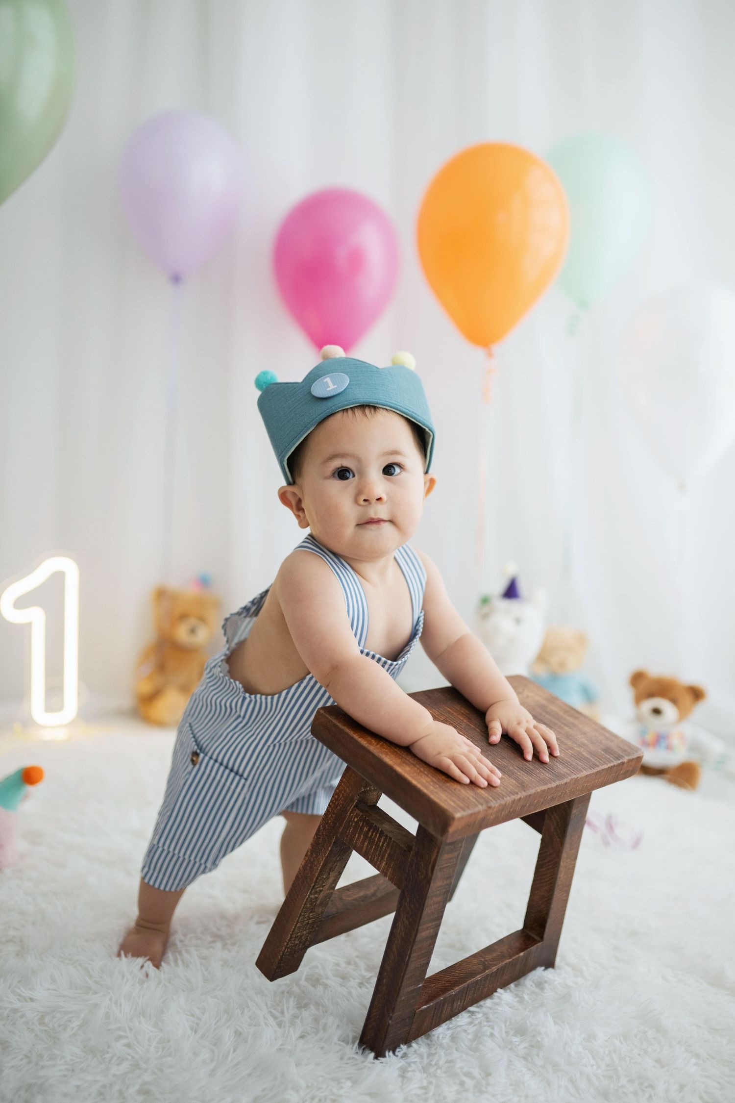 Baby boy in striped overalls and blue bear hat stands at wooden stool surrounded by colorful balloons at first birthday.