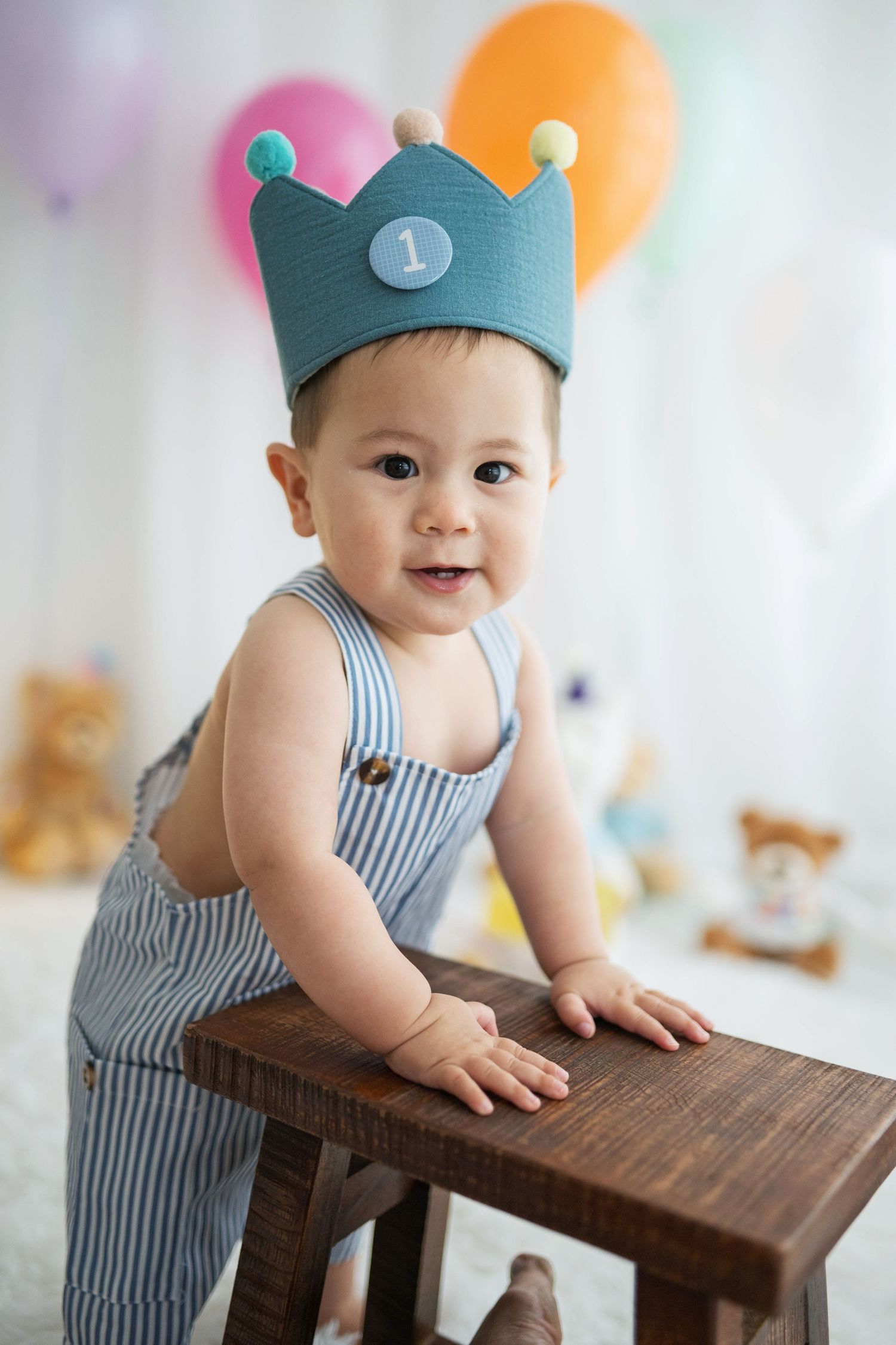 A smiling baby wearing a blue crown with number 1 celebrates their first birthday, leaning on a wooden stool.