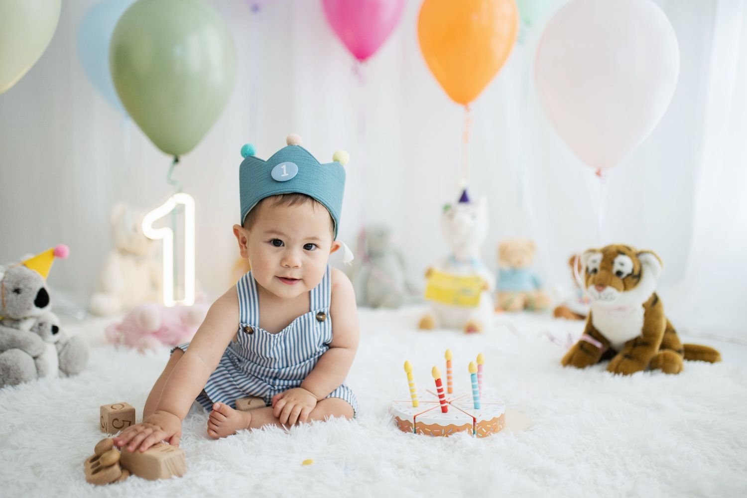 Baby boy in blue crown celebrates first birthday with cake, balloons, and stuffed animals on white fluffy rug.