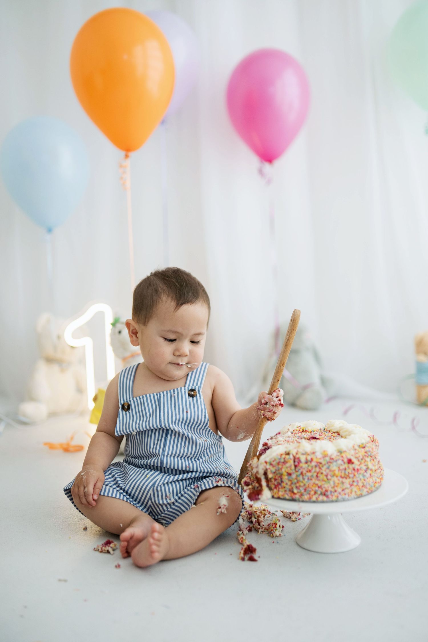 Baby in striped romper smashing first birthday cake surrounded by colorful balloons at a white cake smash photoshoot.