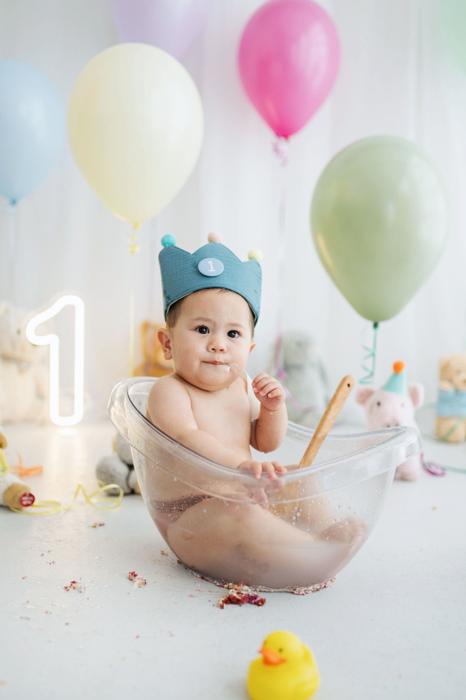 Baby in blue crown sits in bowl surrounded by colorful balloons celebrating first birthday.