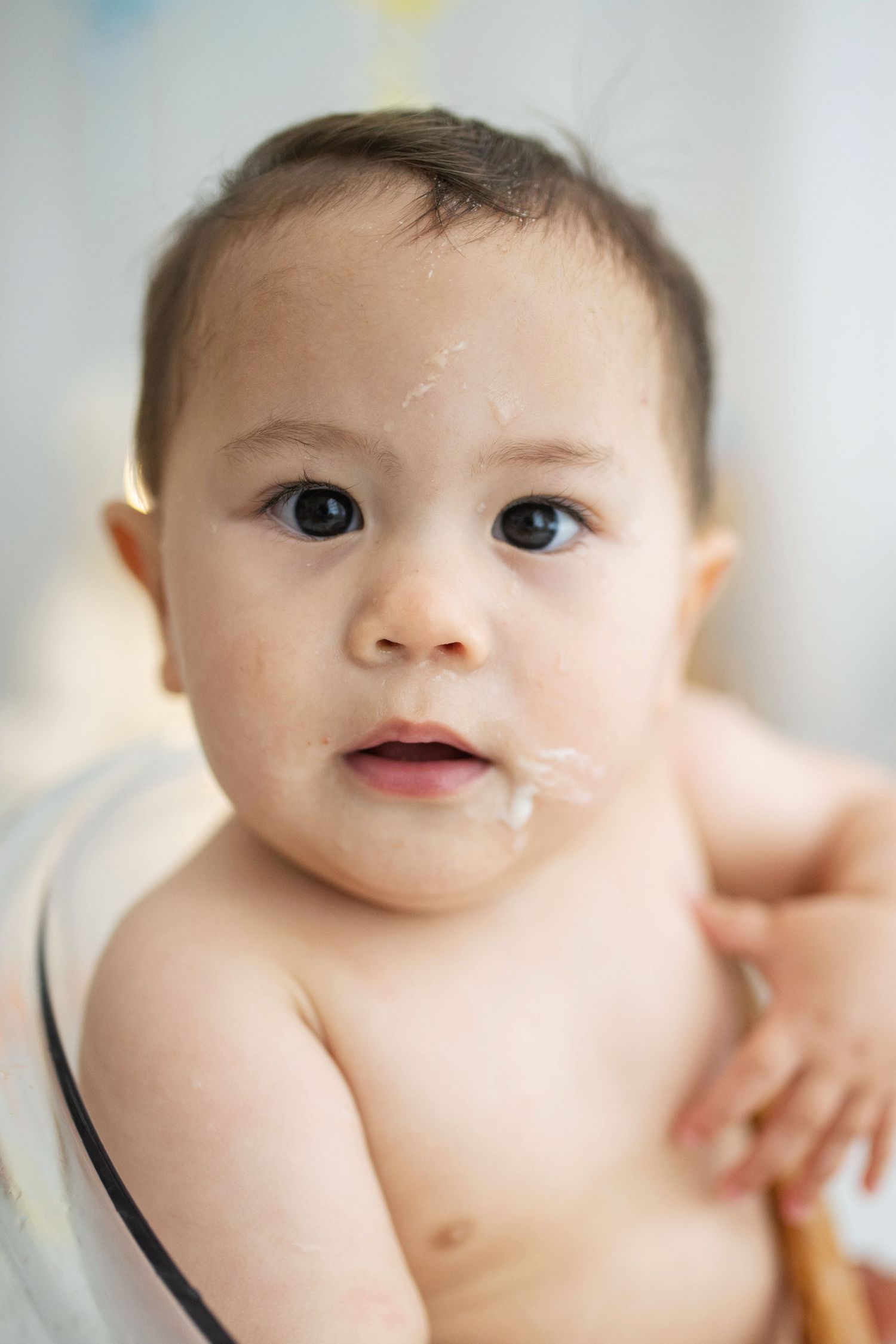 Cute baby with food on face sitting in high chair, looking at camera with big dark eyes.