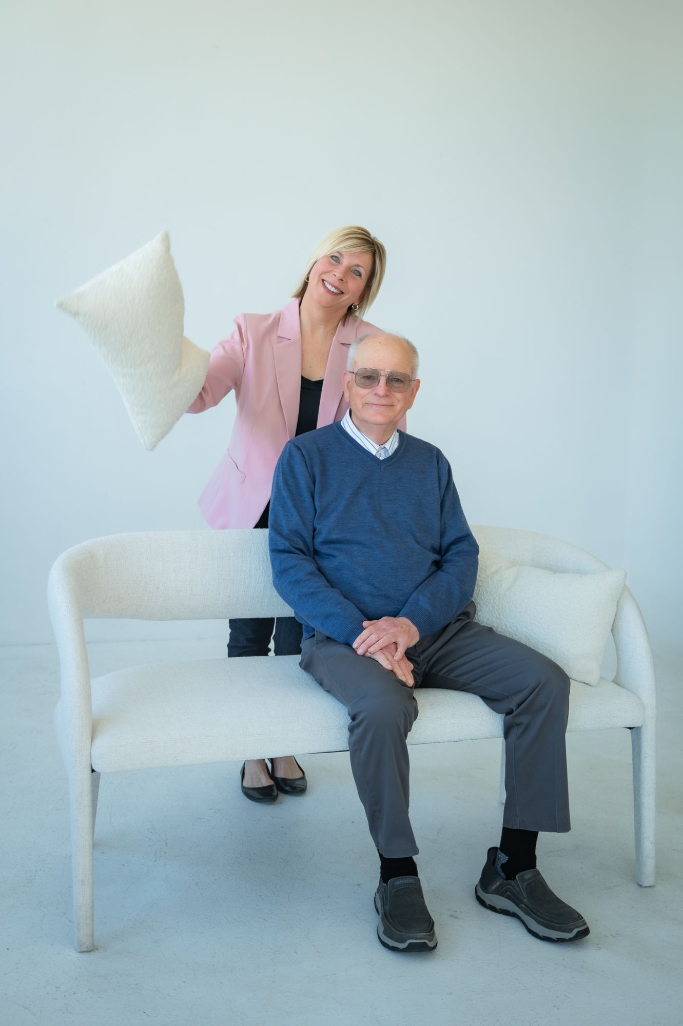 Smiling blonde woman in pink blazer holding pillow stands behind elderly man in blue sweater seated on white bench.