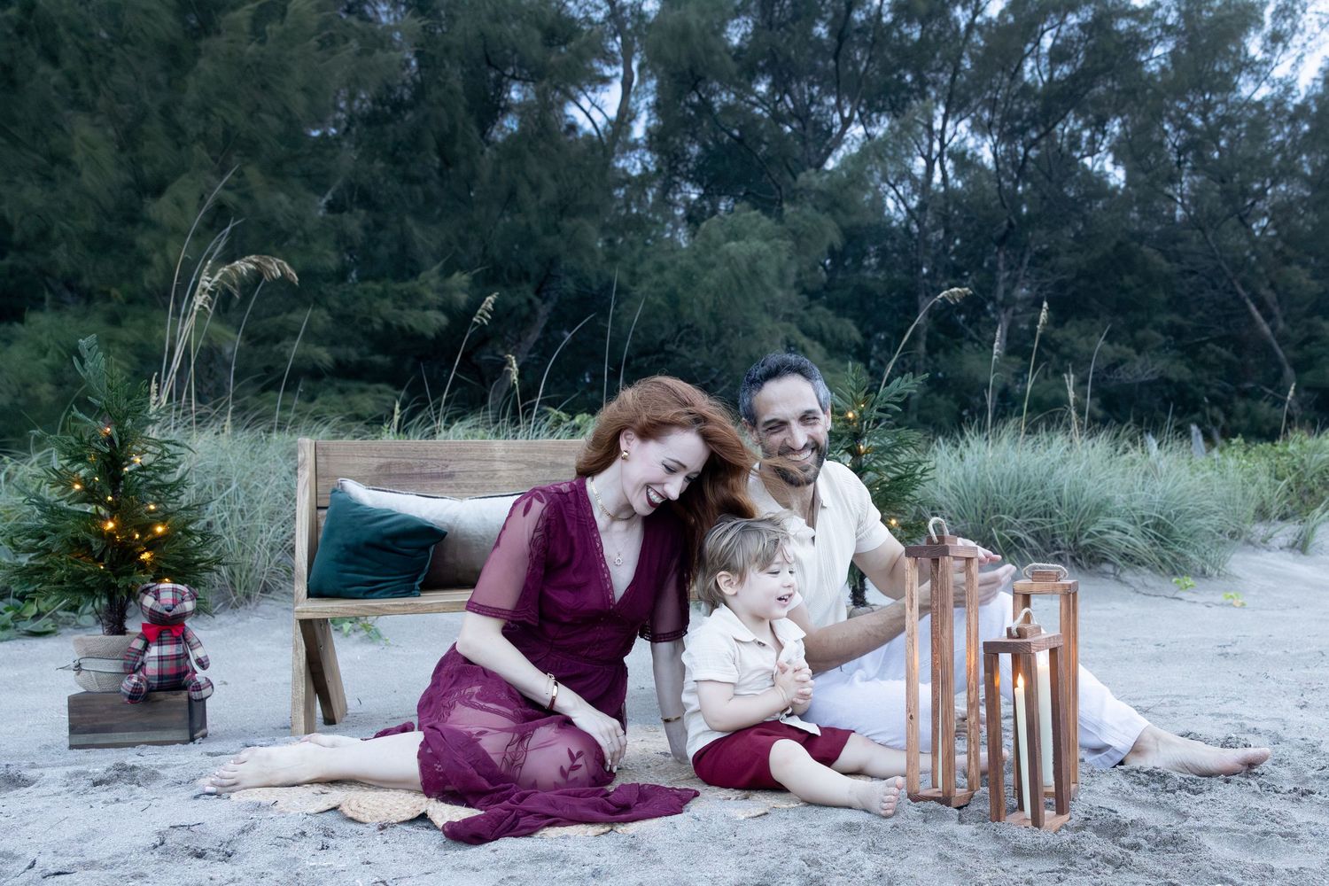 Family of three relaxes on a sandy beach with a wooden bench, a small Christmas tree and a wooden lantern, framed by dunes and trees.