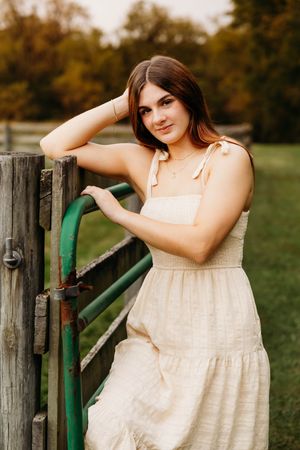 Young woman in cream sundress poses by rustic wooden fence on a farm during golden hour.