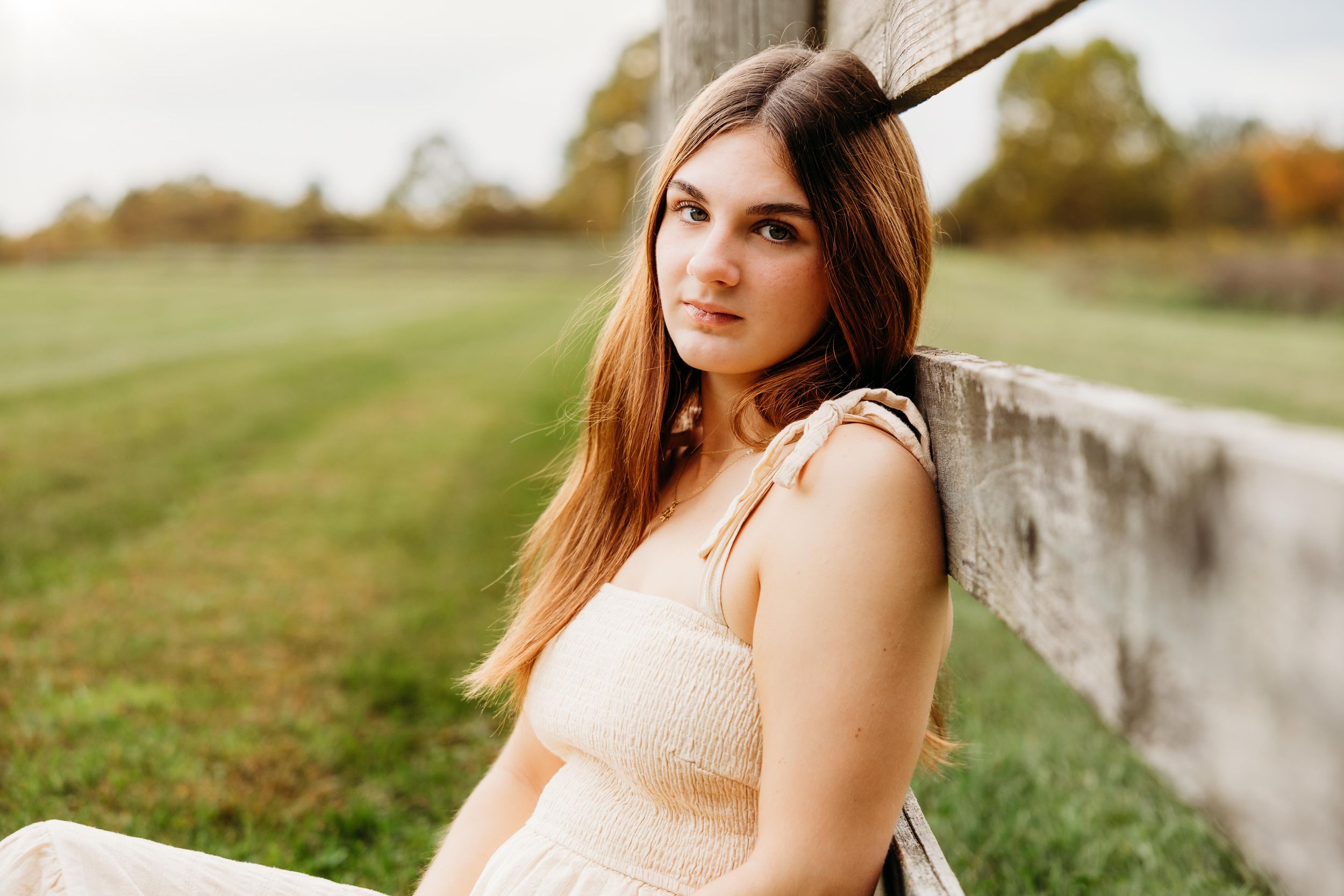 Young woman with long brown hair in a cream dress leaning against a rustic wooden fence in a green field.