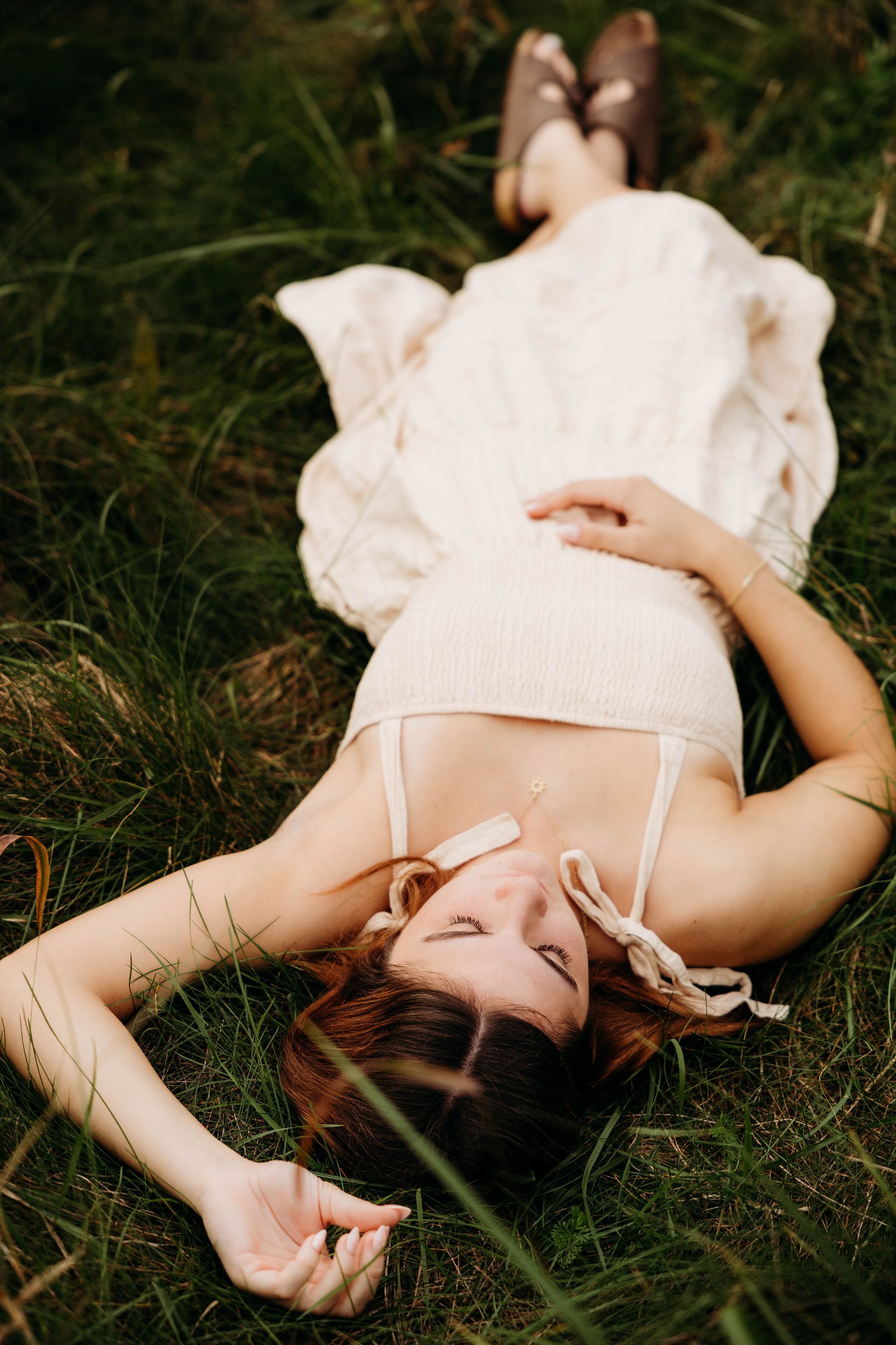 Young woman in white dress lying peacefully on green grass with eyes closed, enjoying nature outdoors.