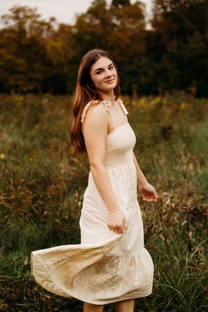 Young woman in cream dress twirling in autumn field, smiling softly with long brown hair flowing.