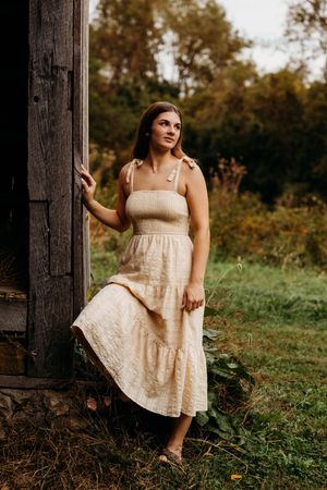 Young woman in a cream tiered midi dress poses gracefully by a rustic wooden barn surrounded by lush greenery.