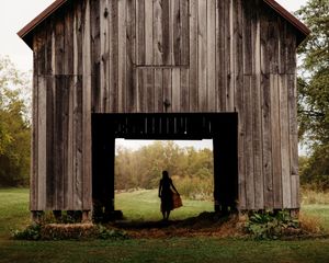 Silhouette of a woman standing in the open doorway of a rustic wooden barn surrounded by lush green fields.