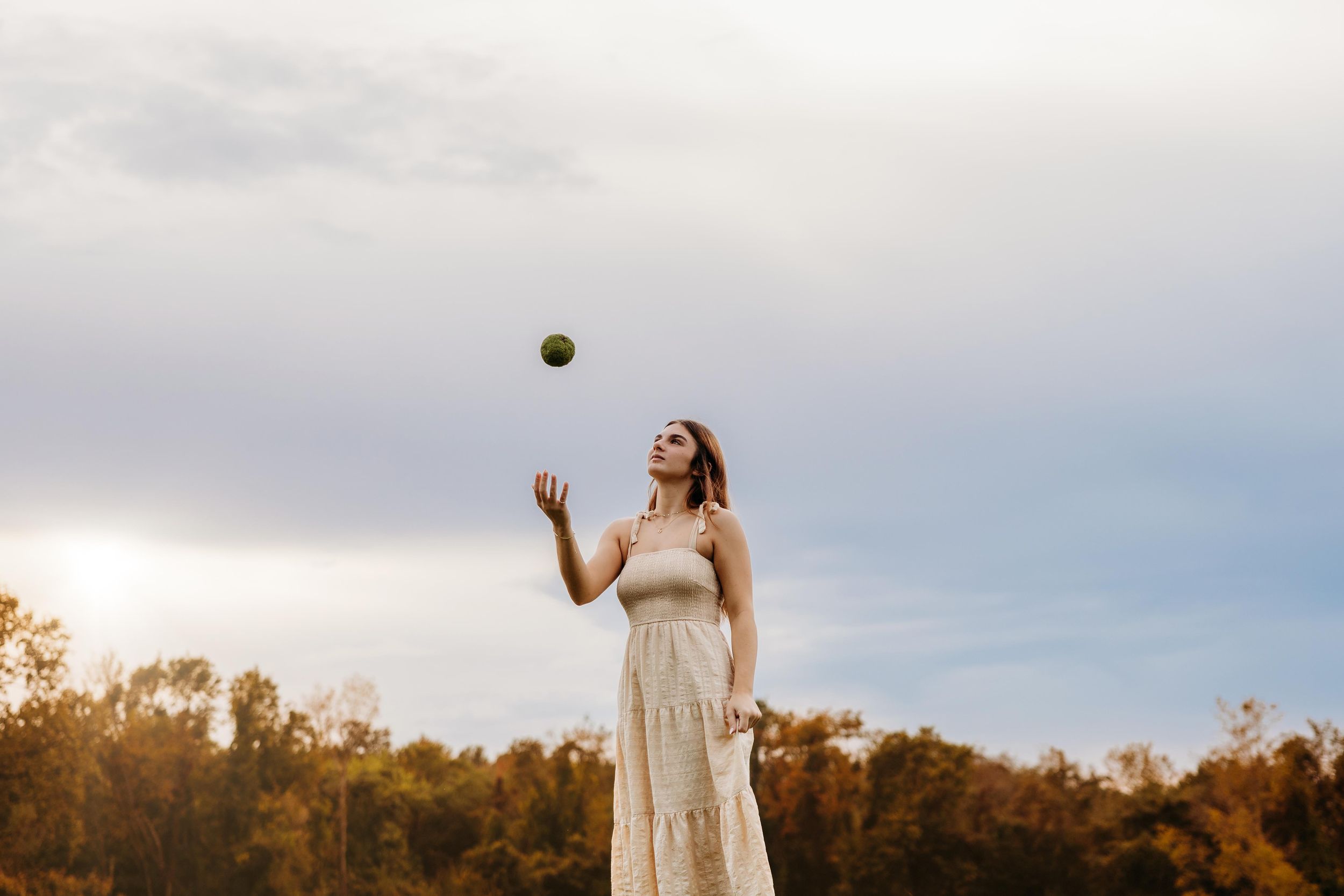 Woman in white dress tossing a green ball into the air outdoors with trees and cloudy sky background.