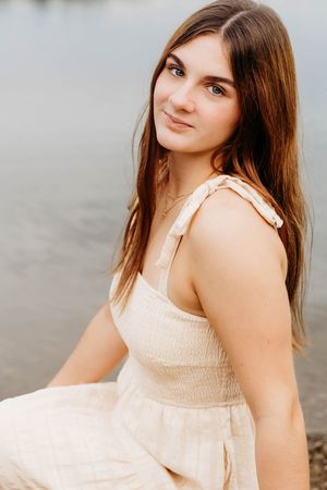 Young woman with long brown hair in a cream smocked sundress sitting by a serene lake, smiling softly.