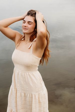 Young woman in cream smocked sundress poses with hands in hair near water, eyes closed, serene expression.