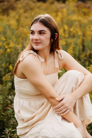 Young woman in a cream smocked dress sitting in a field of yellow wildflowers, smiling softly.