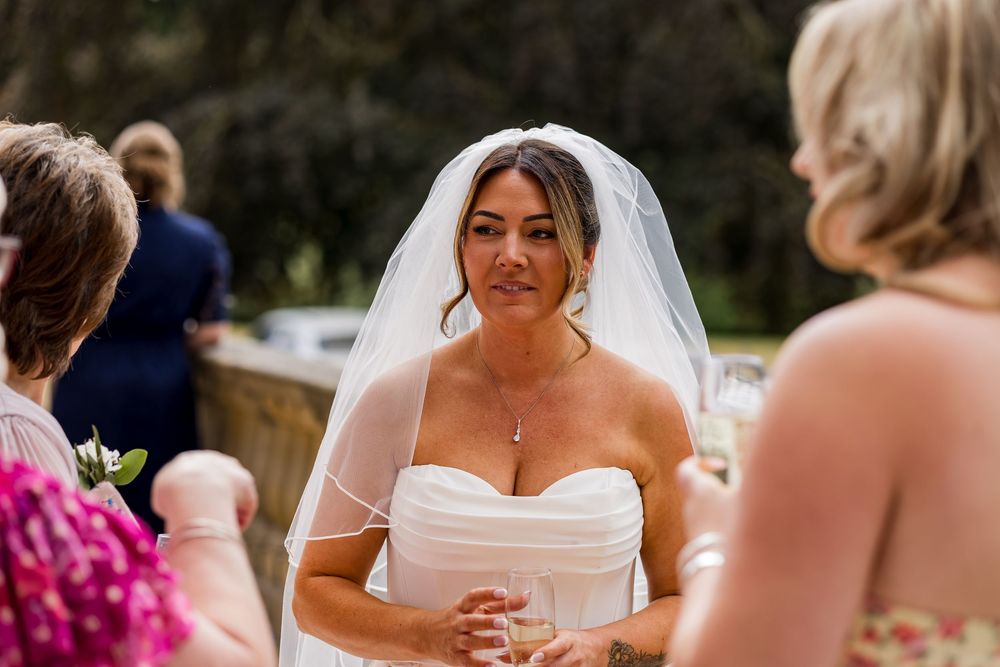 Bride in strapless white gown and veil stands outdoors during wedding ceremony, looking emotional.
