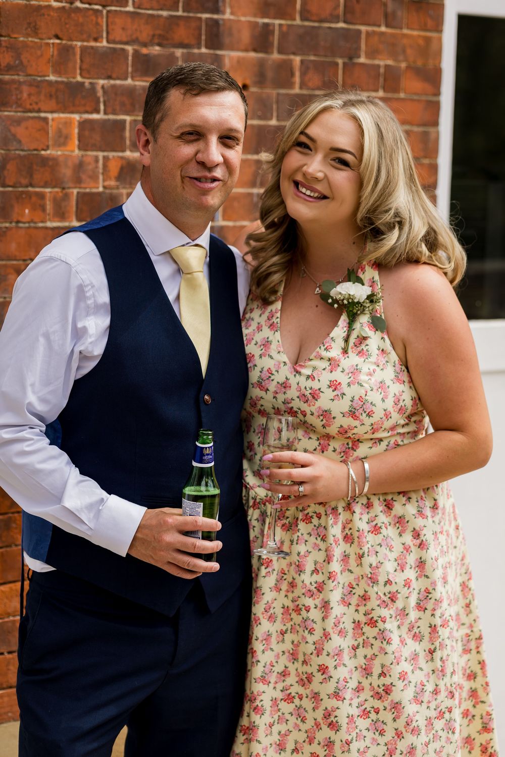 A smiling couple pose together at a wedding, man in navy suit with gold tie, woman in floral dress with corsage.