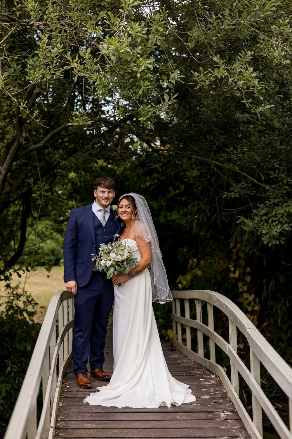 Bride and groom posing on a wooden bridge surrounded by lush green trees on their wedding day.