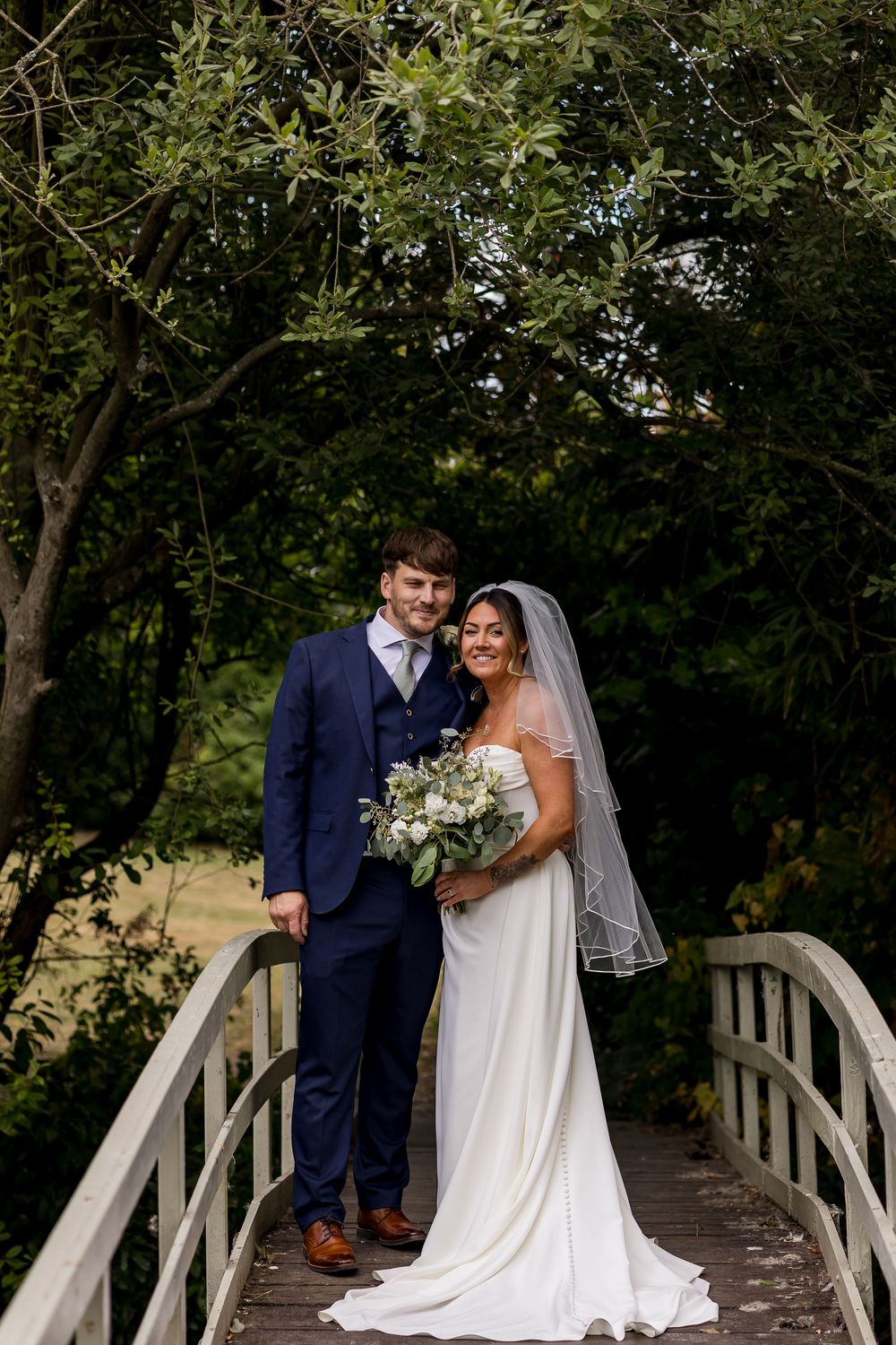 Bride and groom smiling on a wooden bridge surrounded by lush greenery, bride holding white floral bouquet.