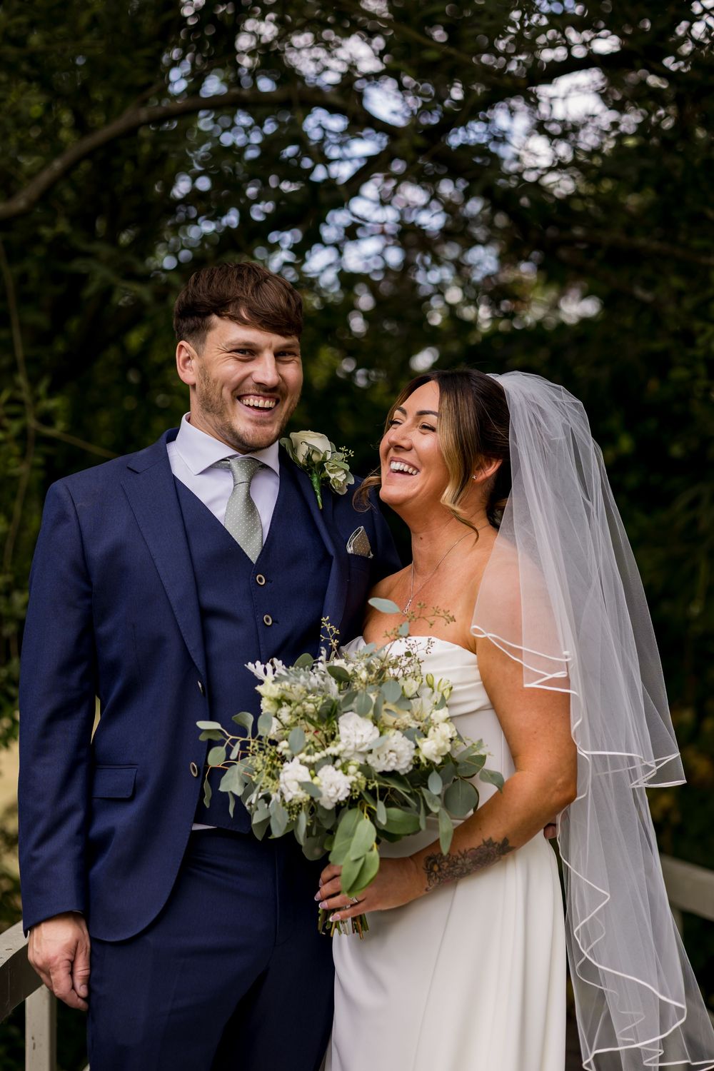 Laughing newlyweds pose outdoors, groom in navy suit, bride in white gown with veil holding white floral bouquet.