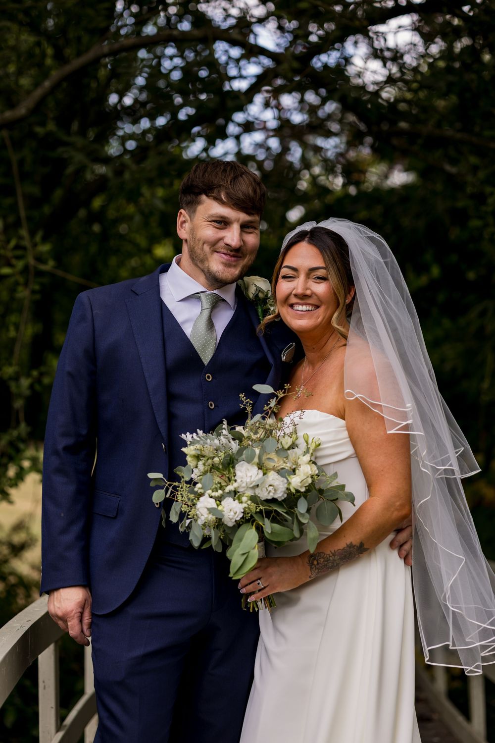 Smiling bride in white gown with veil holding bouquet stands with groom in navy suit outdoors.