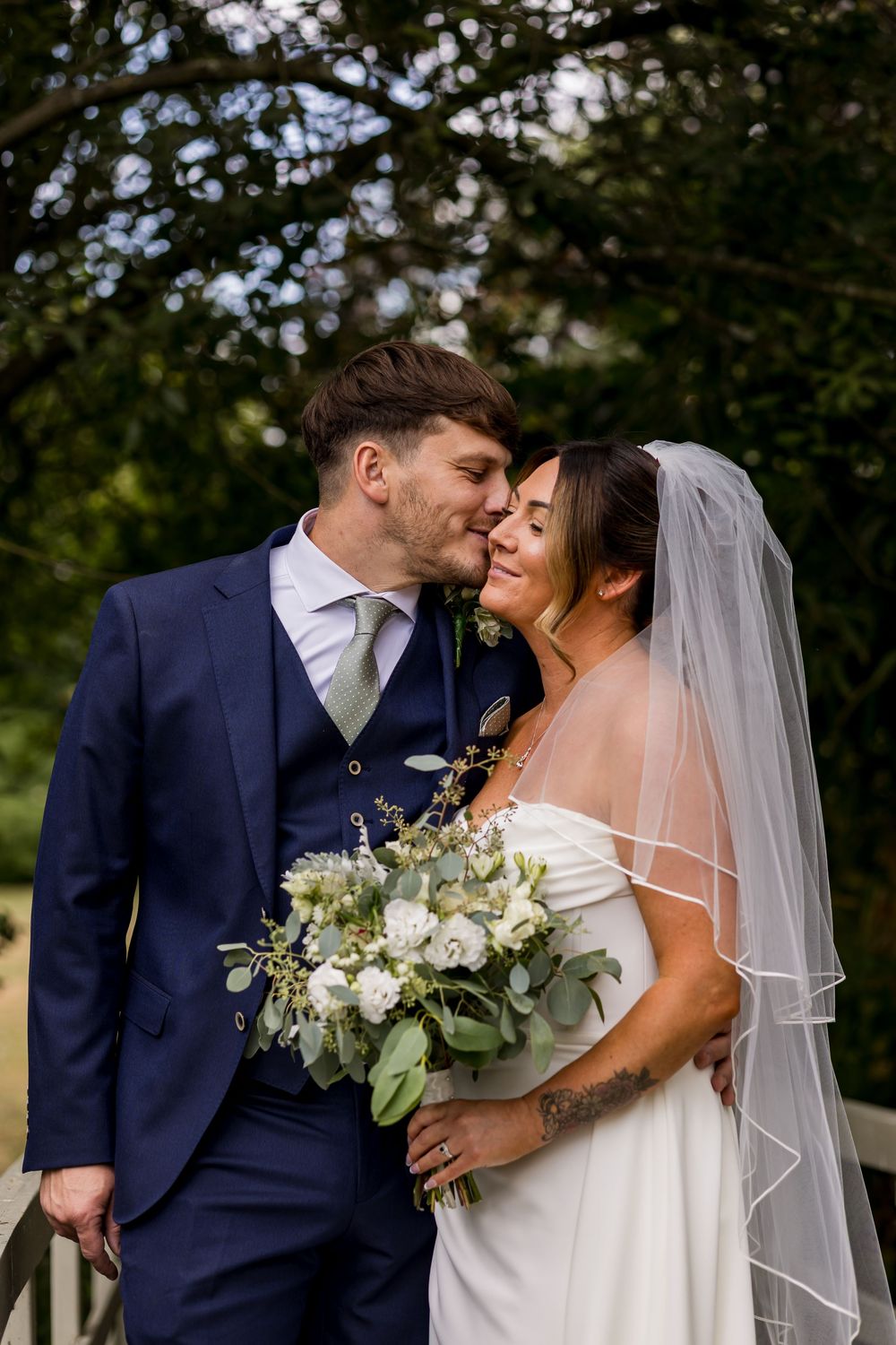 Bride and groom share a romantic kiss outdoors, she holds white floral bouquet wearing veil, he wears navy suit.