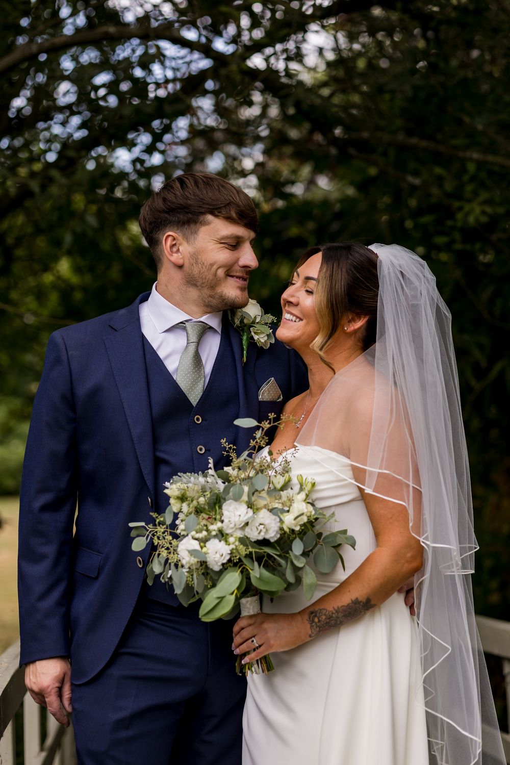 Bride and groom share a romantic moment, she holds white floral bouquet wearing veil, he wears navy three-piece suit.