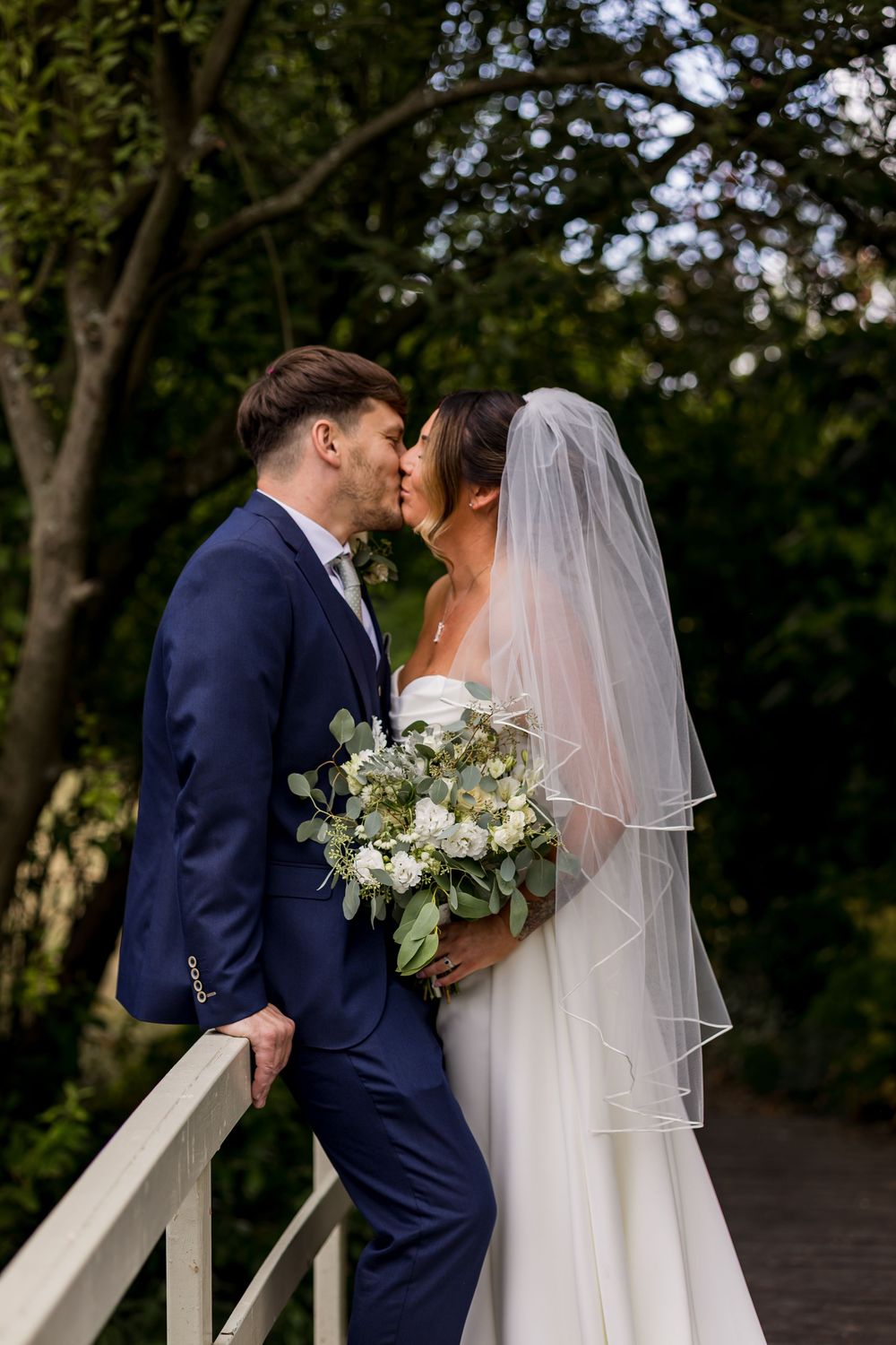 Bride and groom share a romantic kiss on a garden bridge, bride holding white floral bouquet in elegant wedding attire.