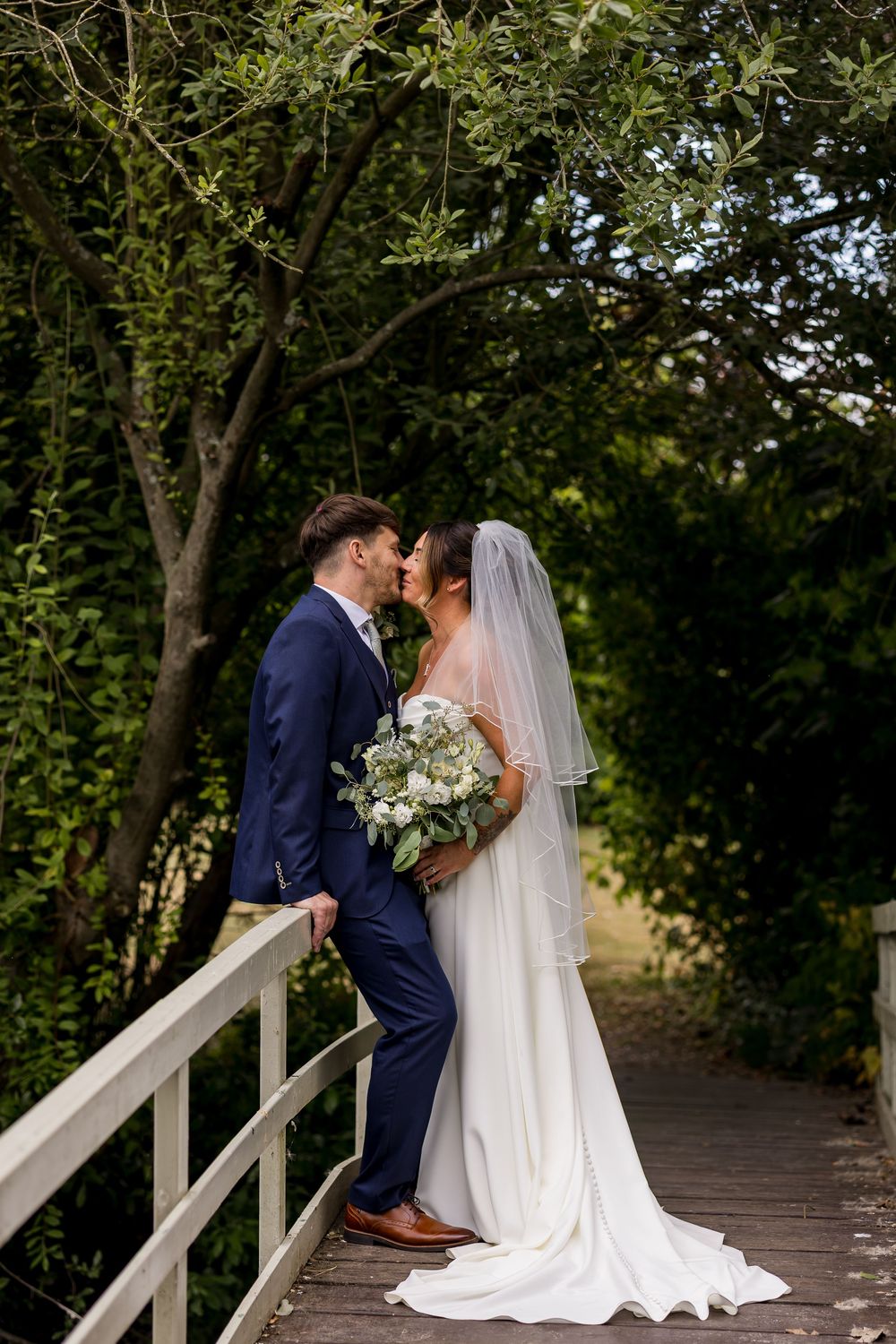 Bride and groom share a romantic kiss on a wooden bridge surrounded by lush green trees, bride holding white floral bouquet.