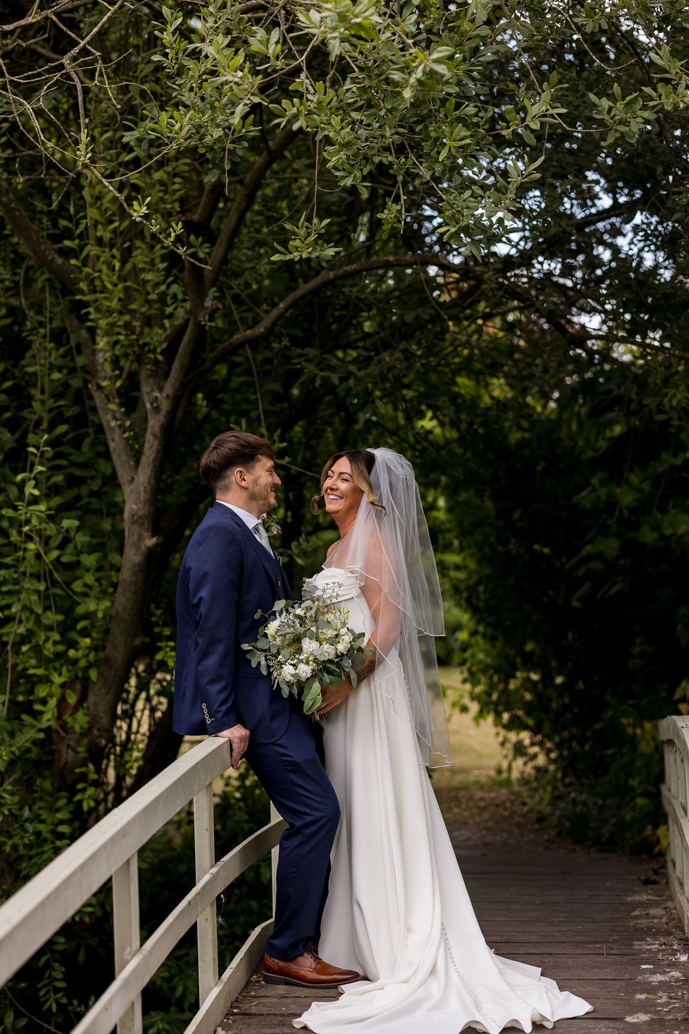 A bride and groom share a romantic moment on a wooden bridge surrounded by lush green trees on their wedding day.