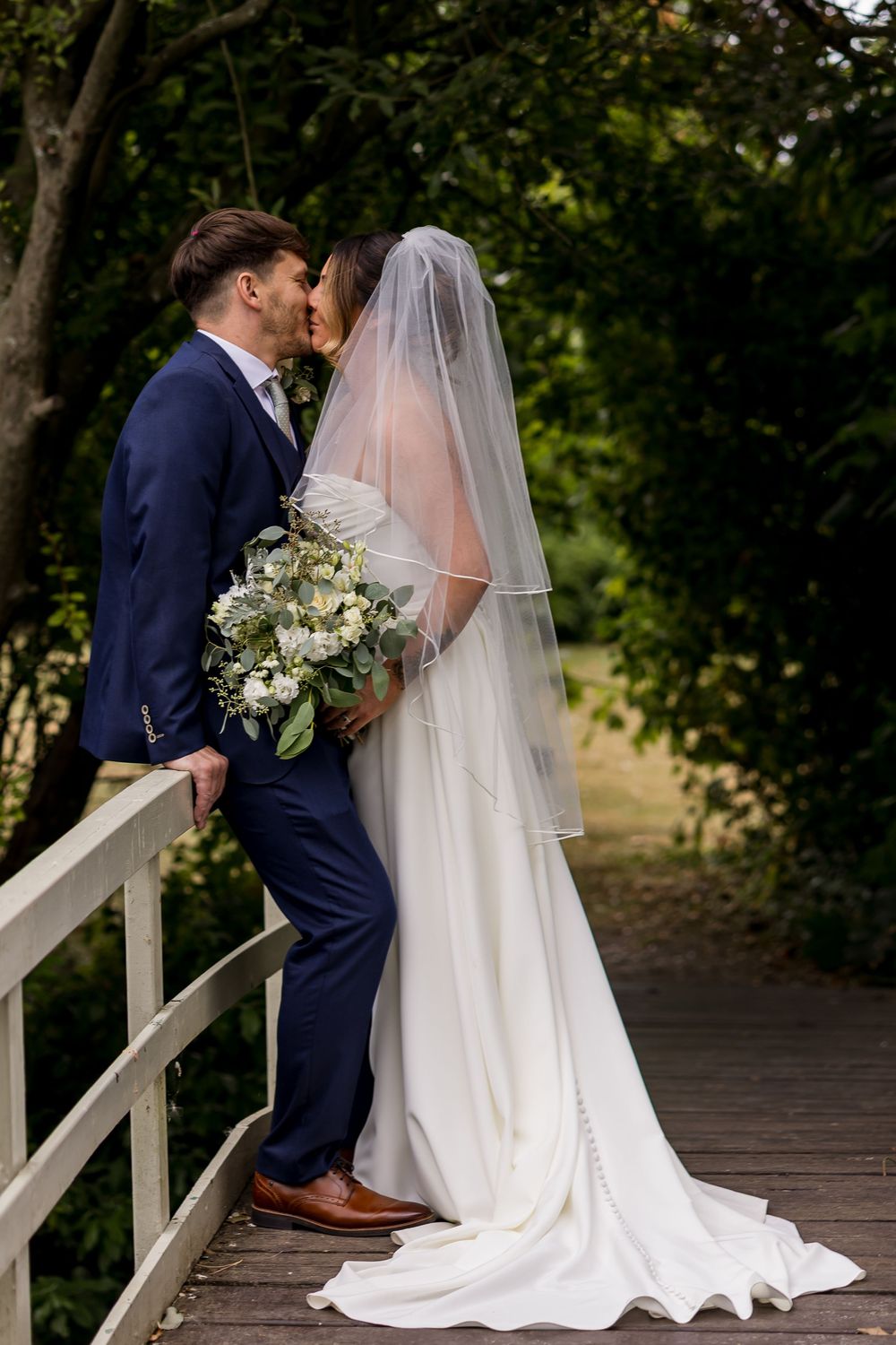 Bride and groom sharing a kiss on a wooden bridge, bride in flowing white gown with veil holding bouquet.