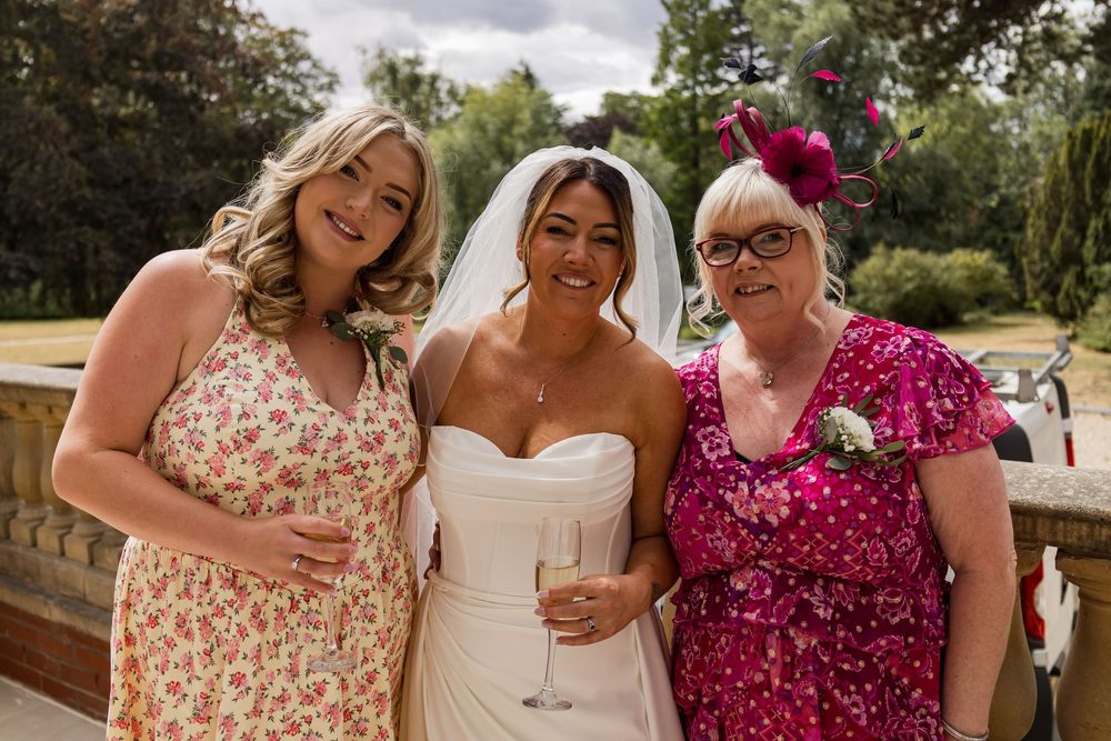 Bride in white strapless gown poses with two women in floral dresses at an outdoor wedding celebration.