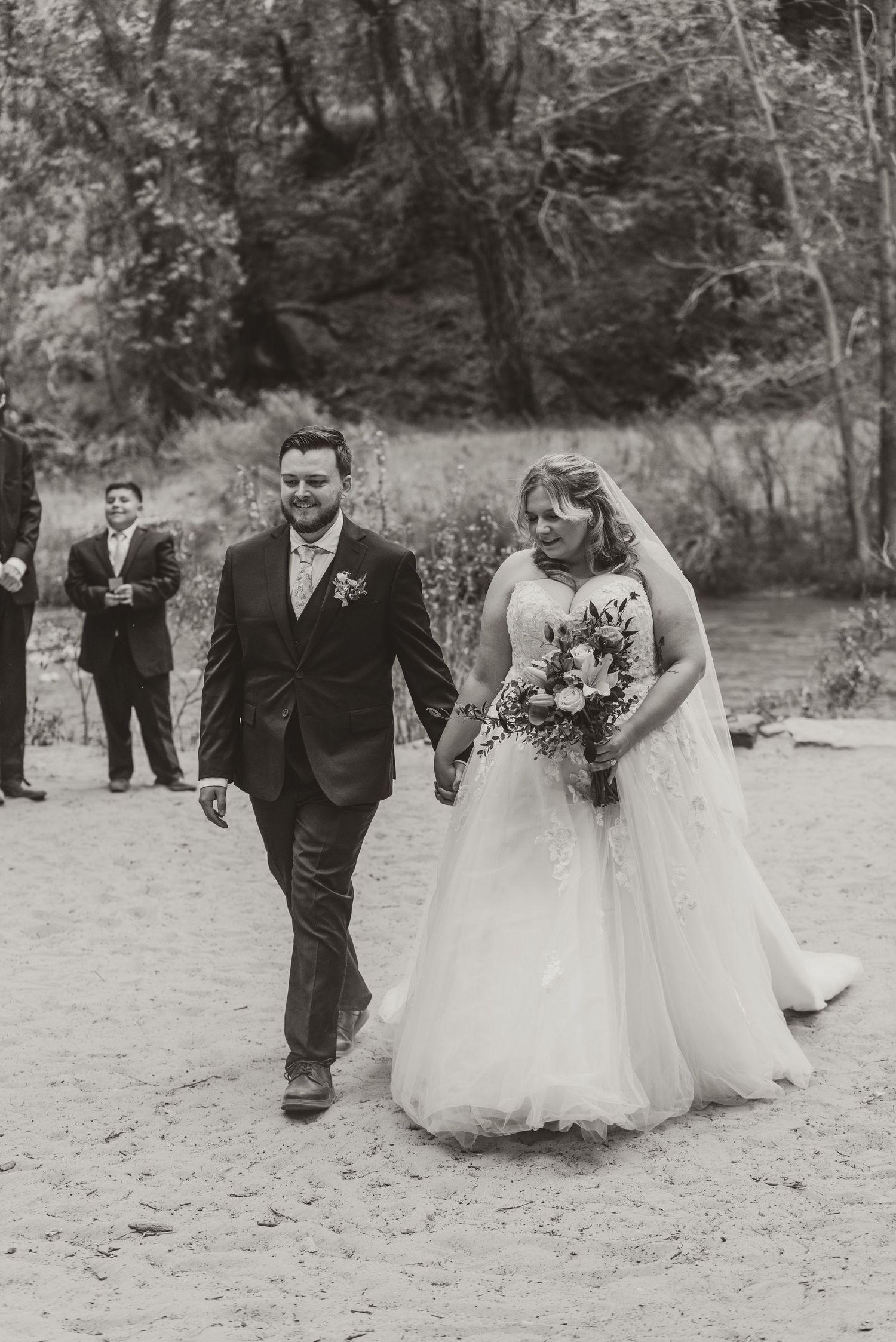 Black and white photo of a smiling bride and groom holding hands, walking outdoors after their wedding ceremony.