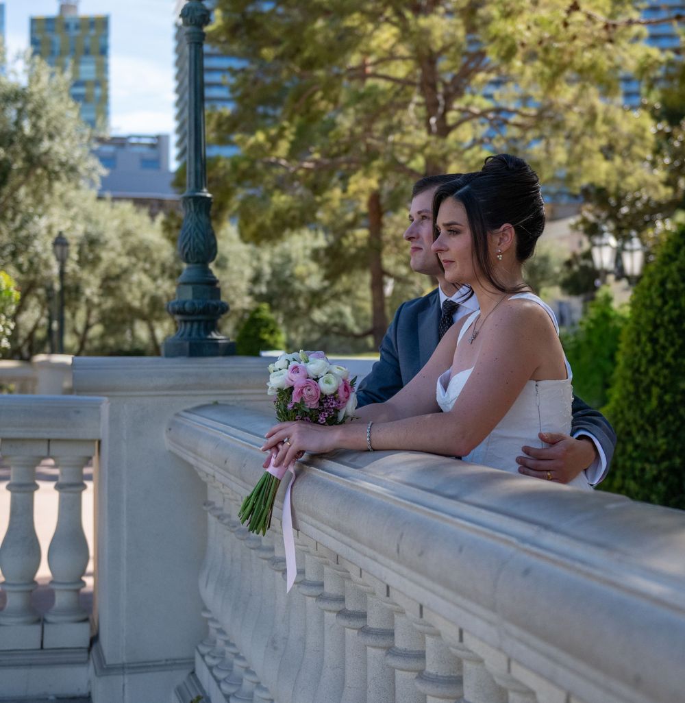 Bride holding a pink bouquet and groom lean on a stone balustrade in a lush garden setting.