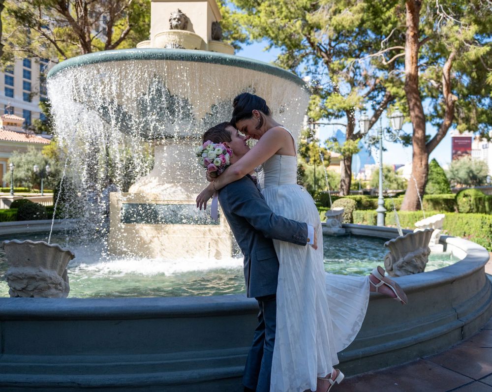 Newlyweds embrace in a romantic kiss in front of an elegant fountain surrounded by lush trees.