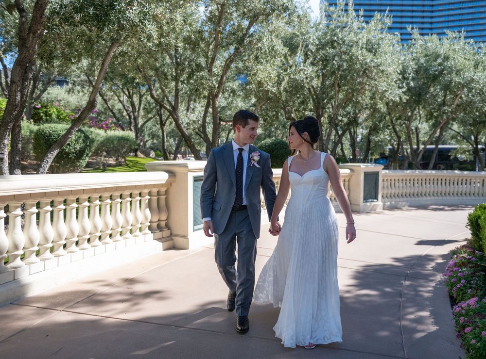 Bride and groom walking hand in hand along an elegant balustrade pathway surrounded by lush olive trees on their wedding day.
