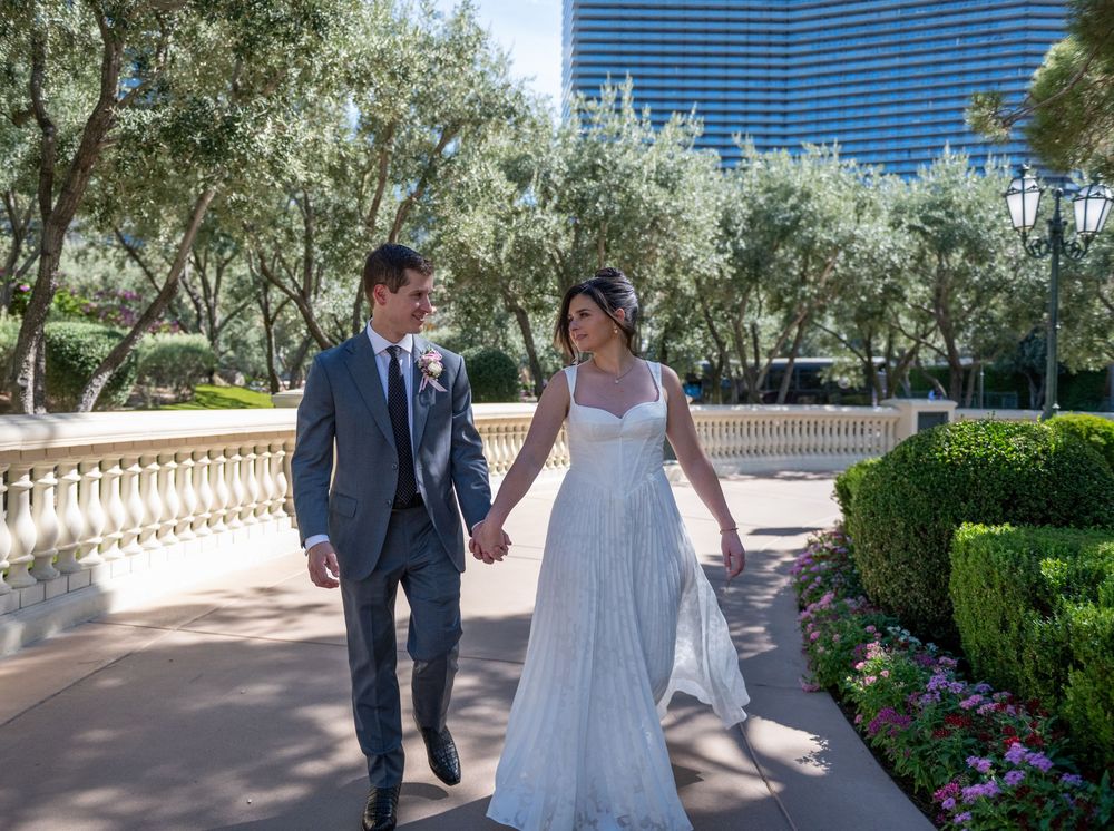 A bride and groom walk hand in hand through lush garden grounds with a modern Las Vegas hotel tower in the background.