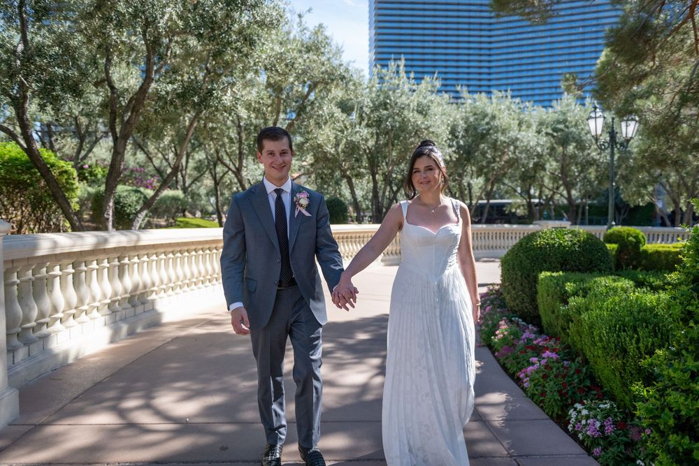 A bride and groom hold hands walking along a garden path in Las Vegas, with lush greenery and a modern hotel tower behind them.