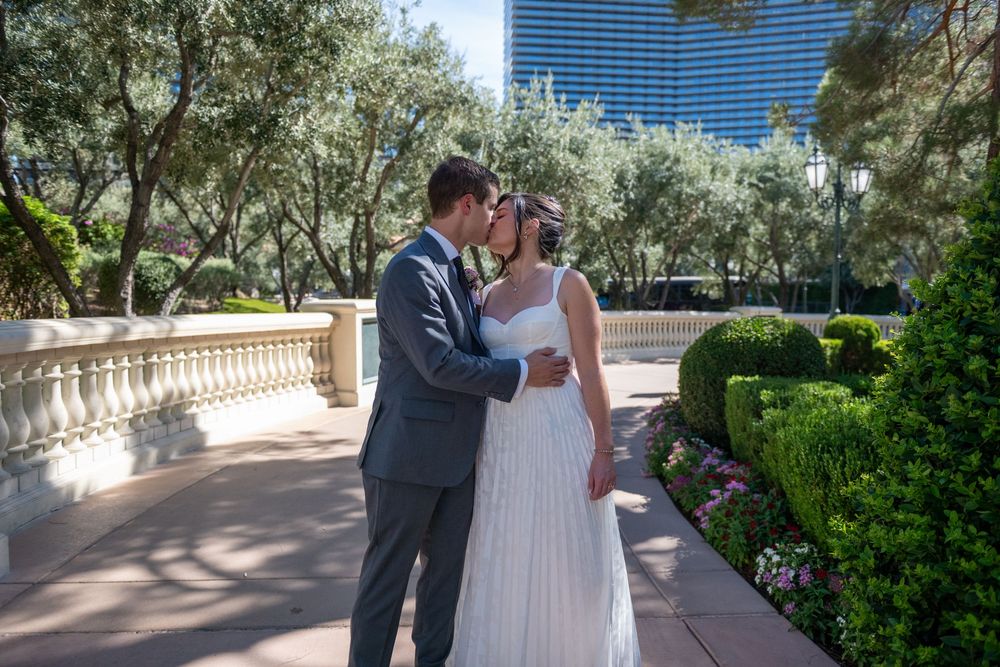 Bride and groom share a romantic moment in a lush garden with tall trees and colorful flowers, Las Vegas skyline in background.