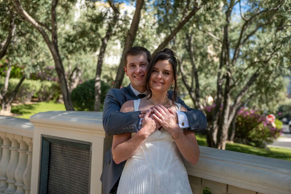 A happy couple in wedding attire embraces on a balcony surrounded by lush green olive trees.