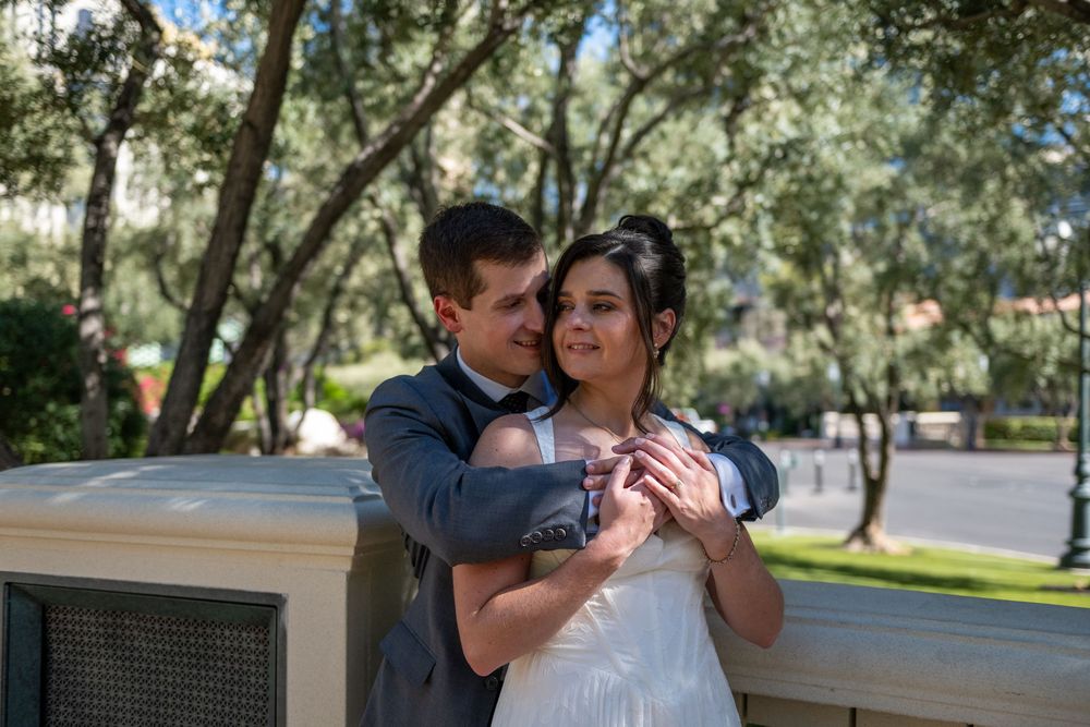 A groom in a gray suit embraces his bride in a white dress outdoors, surrounded by lush green trees on their wedding day.
