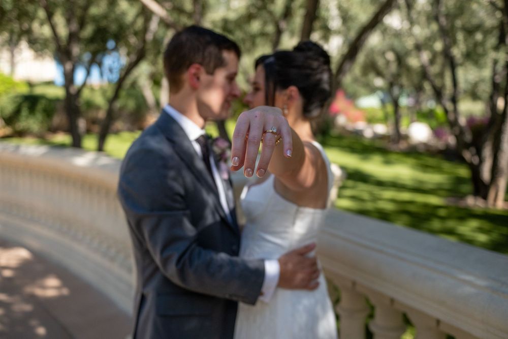 Newlywed couple showing off wedding rings outdoors near white balustrade with lush green trees in background.
