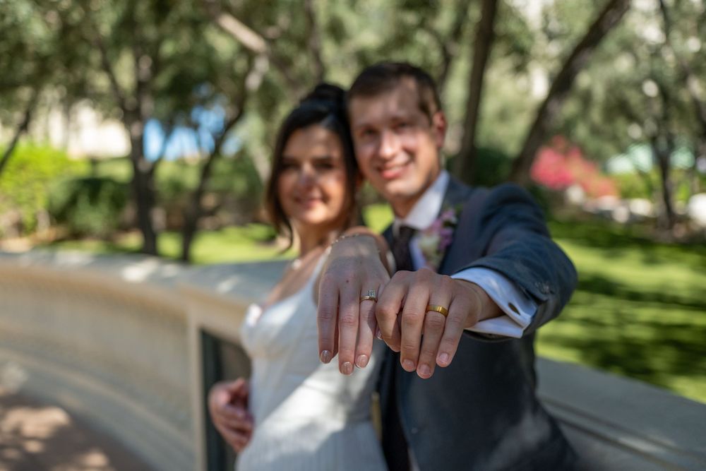 Newlywed couple showing off wedding rings outdoors, bride in white dress and groom in suit, green trees background.