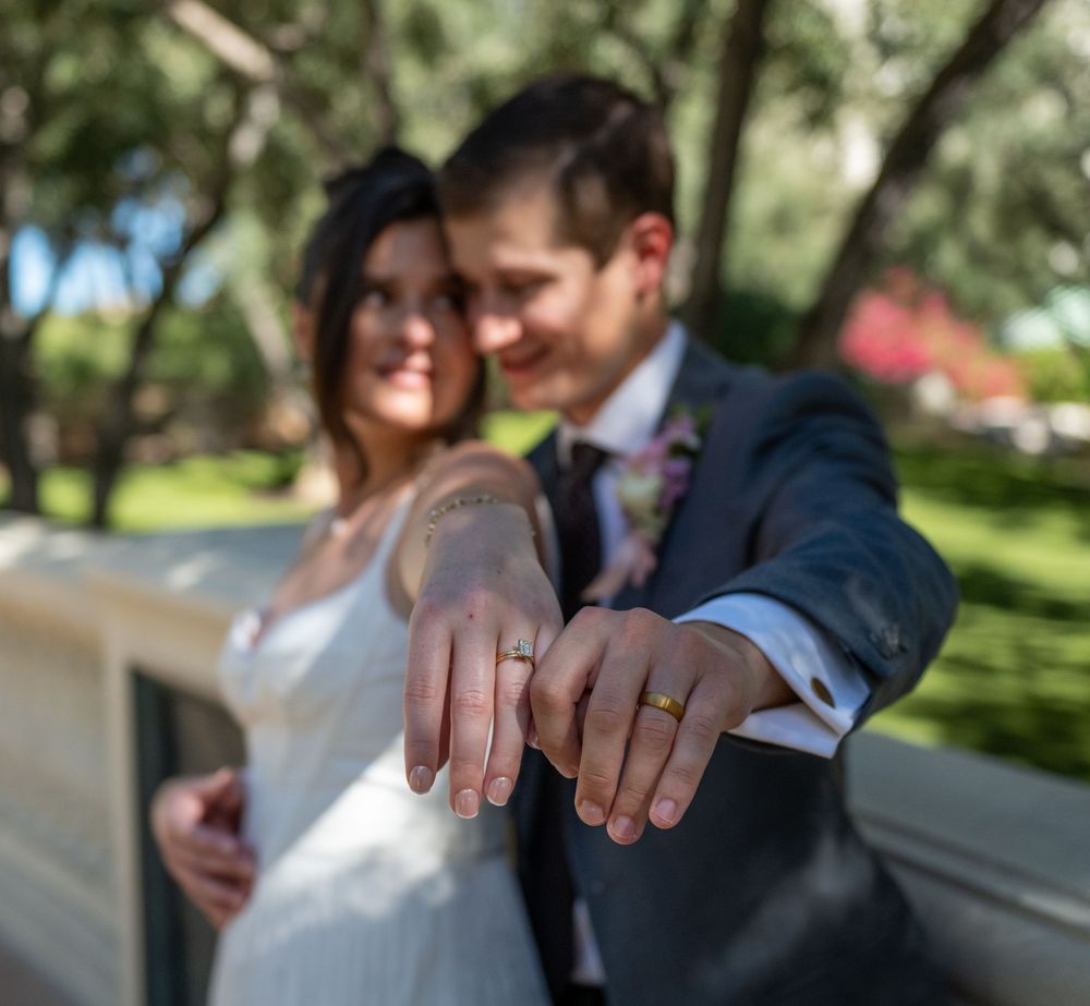 Newlywed couple showing off wedding rings outdoors, bride in white dress and groom in suit, smiling together.