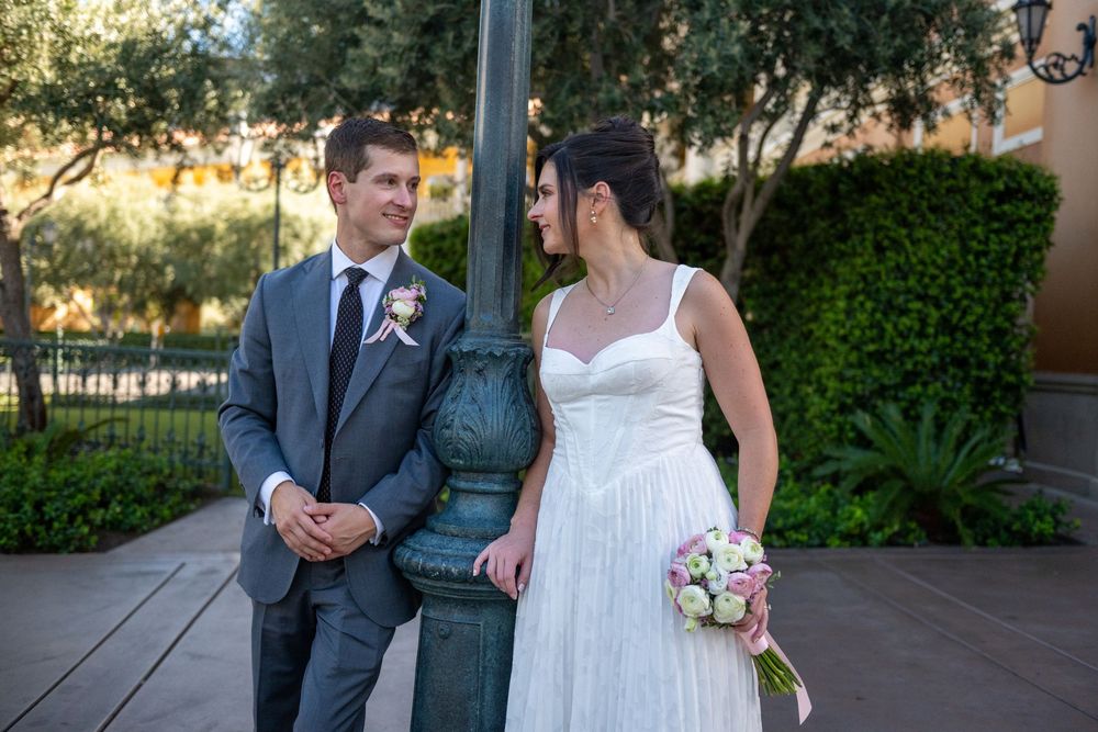 Bride and groom stand by a vintage lamppost, she holds a pink bouquet, wearing a white gown, he in a gray suit.