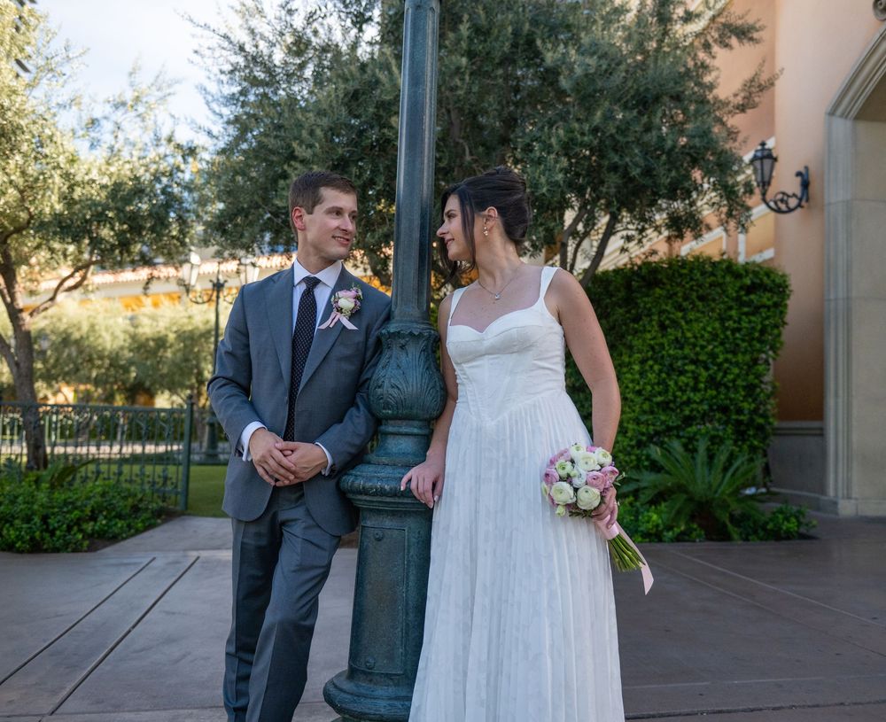 Bride and groom in wedding attire standing by a green lamppost outdoors, exchanging glances on their wedding day.