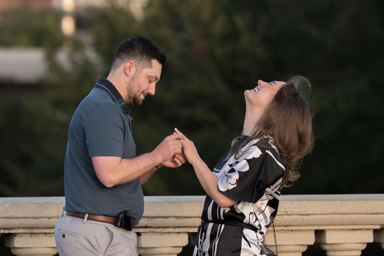 A man in a gray polo holds a woman's hand as she tilts her head back laughing, in a joyful moment on a stone balustrade outdoors.
