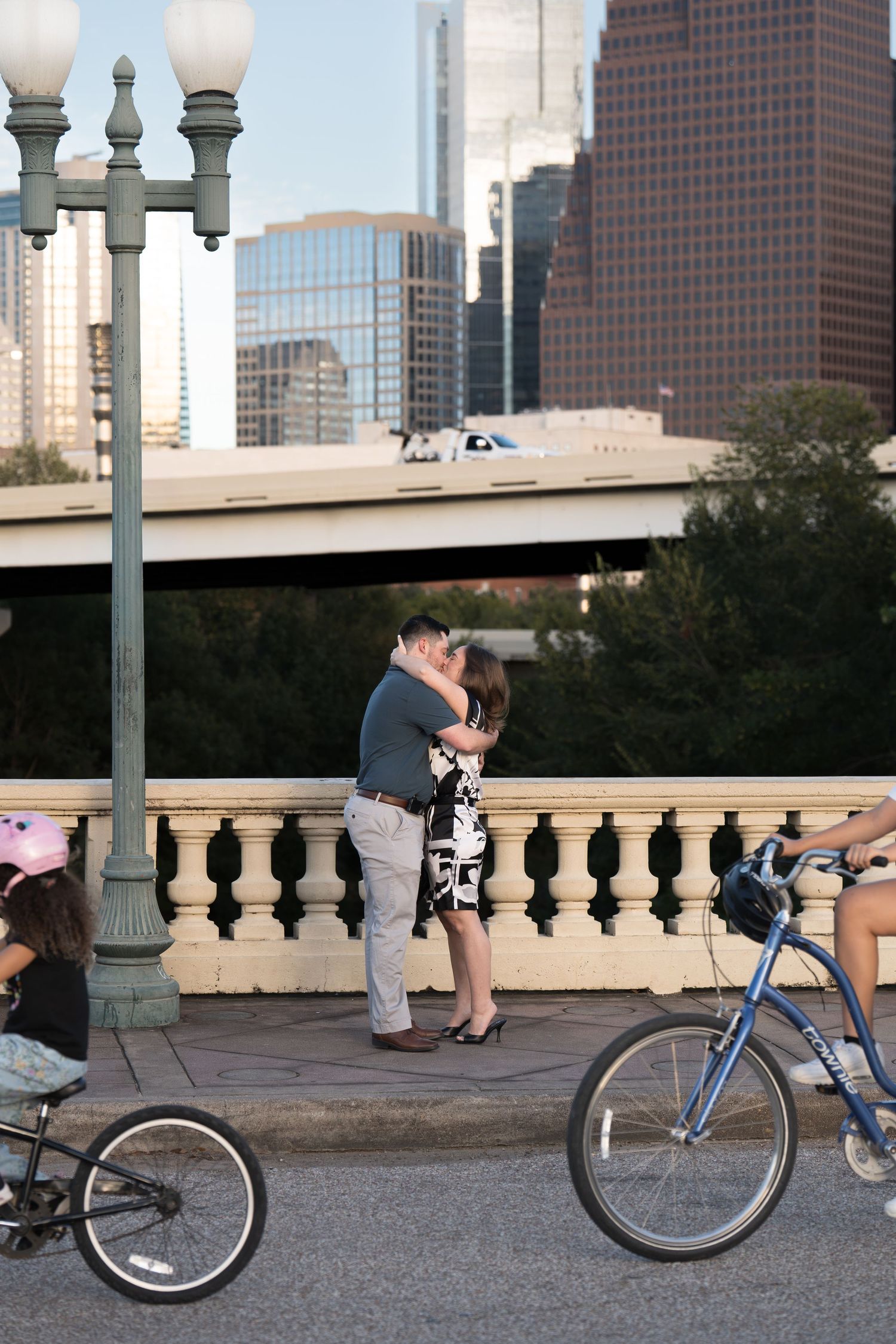 A couple embraces on a city bridge with a vintage lamppost, cyclists passing by, and downtown skyscrapers behind them.