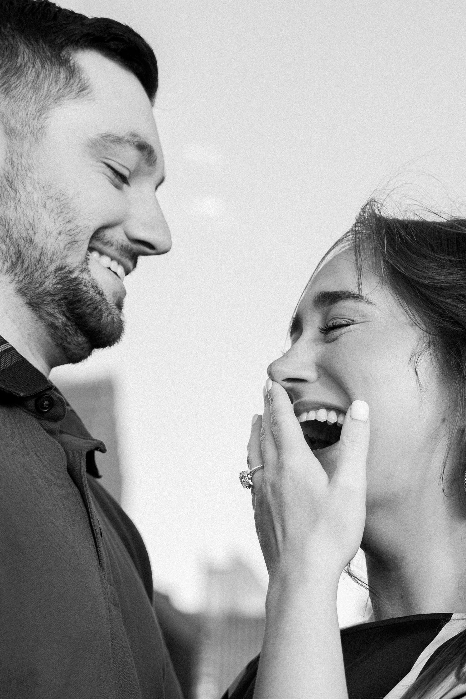 Black and white photo of a smiling bearded man and a laughing woman covering her mouth, showing a sparkling engagement ring.